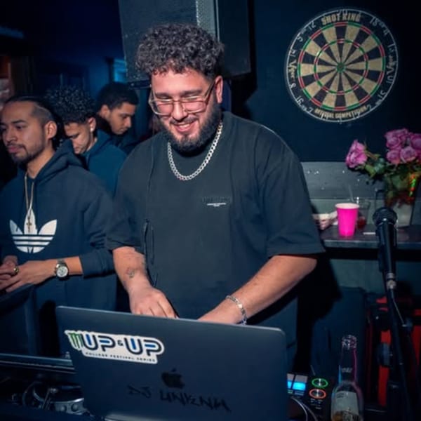 A DJ with glasses and a beard smiles while mixing music on a laptop at a dark venue.